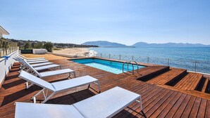 Un espace aménagé au bord de la piscine, avec des chaises longues et une terrasse en bois offrant une vue sur la plage et la mer.