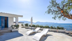 Une terrasse sur le toit avec des chaises longues blanches, un bâtiment blanc avec une porte bleue, et une vue sur la mer et les montagnes.
