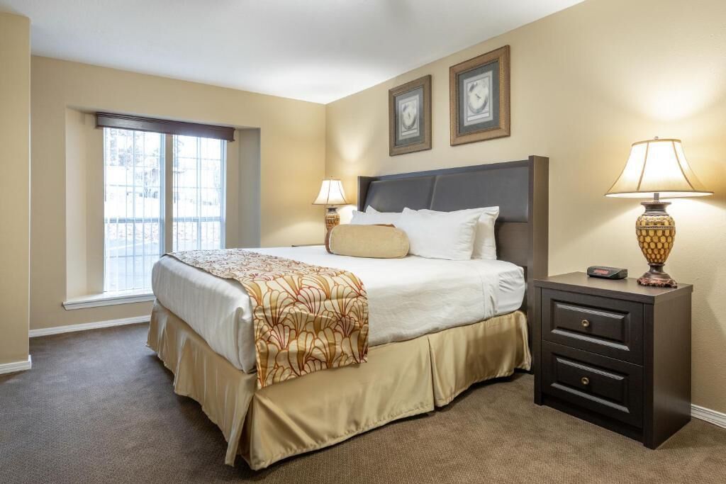 A hotel room with a large bed, two bedside lamps, a nightstand, and a window with blinds.