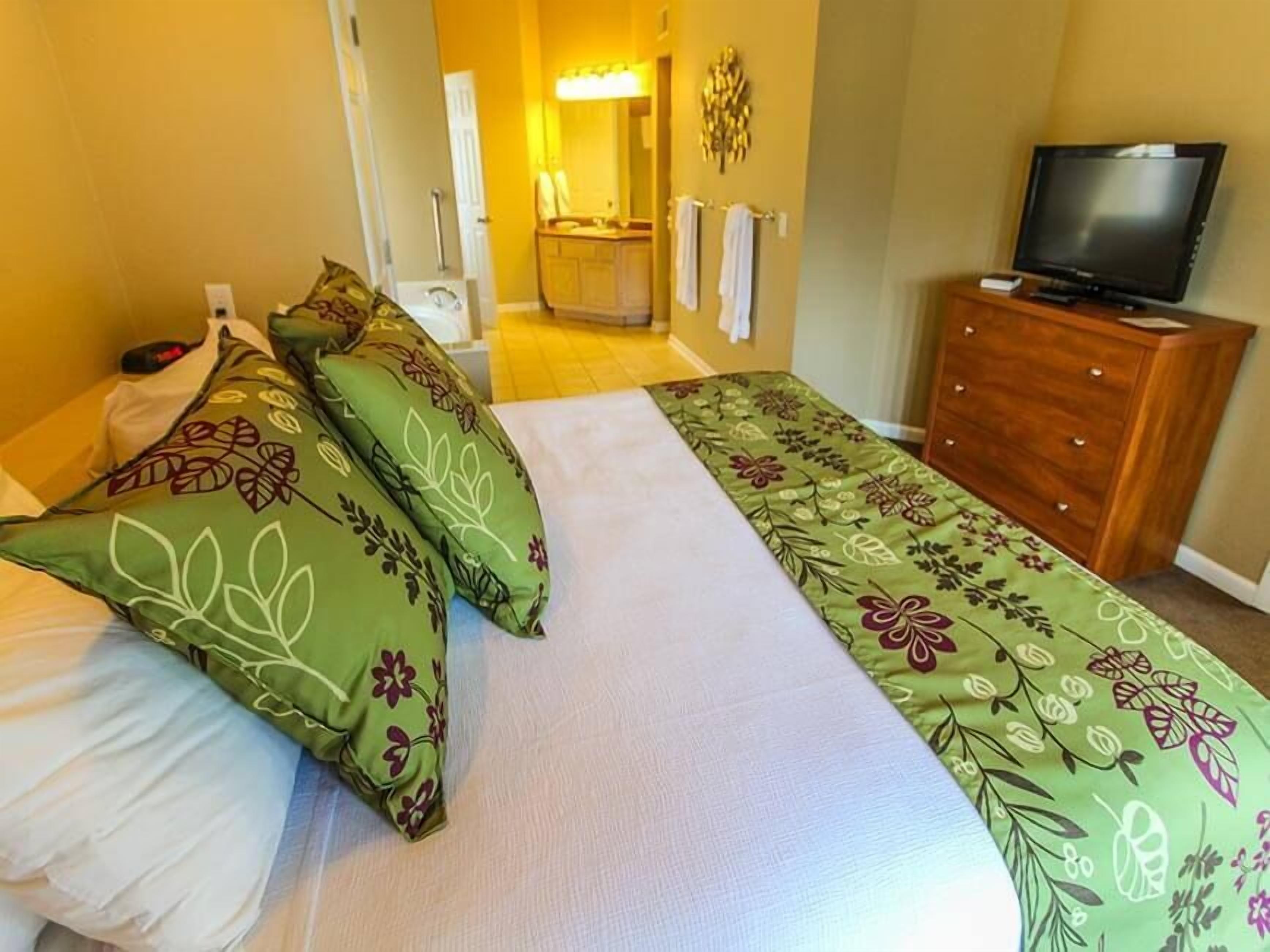 A hotel room with a bed, a TV, a wooden dresser, and a bathroom visible in the background.