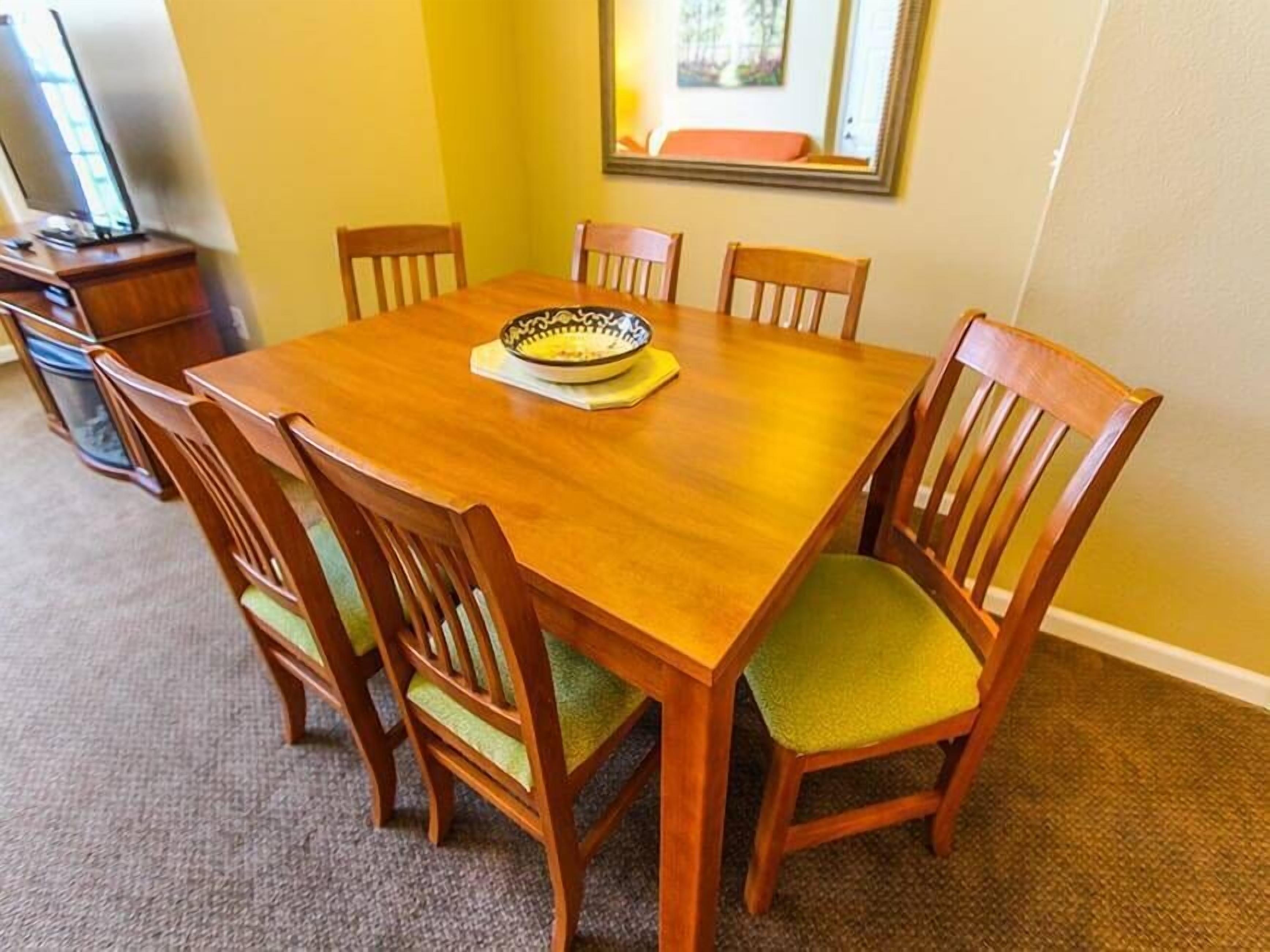 A dining area with a wooden table and six chairs, a bowl on the table, and a framed picture on the wall.