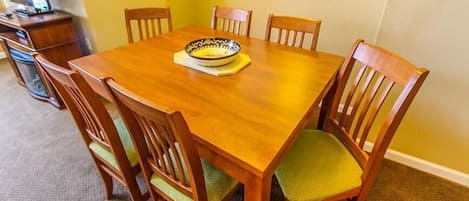 A dining area with a wooden table and six chairs, a bowl on the table, and a framed picture on the wall.