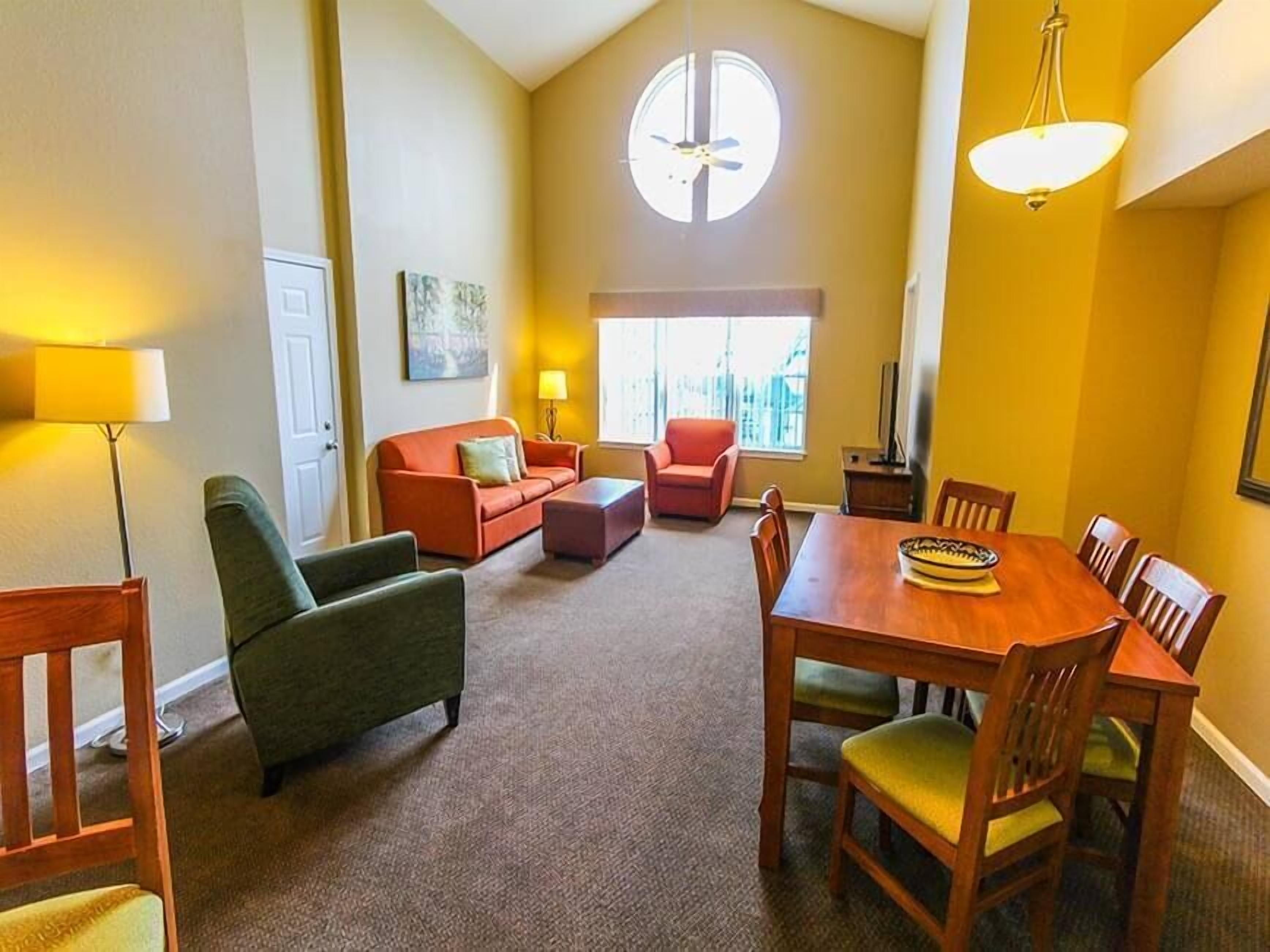 A spacious living room with a high ceiling, a dining area with wooden furniture, and a large window providing natural light.