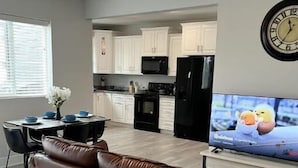 Uma sala de estar com sofá de couro marrom, uma mesa de jantar com pratos azuis, uma cozinha com armários brancos, uma geladeira preta, um ventilador de teto e uma televisão exibindo um desenho animado.