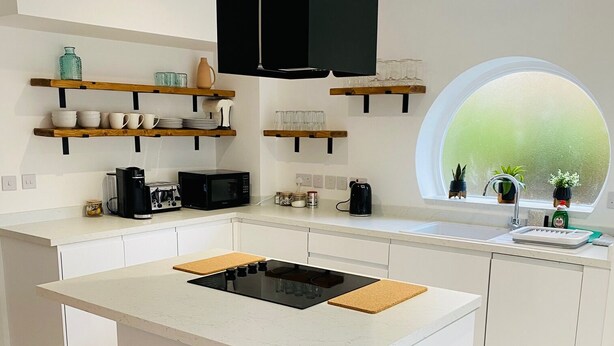 A modern kitchen with a central island, bar stools, a black trash bin, and wooden shelves with kitchenware.