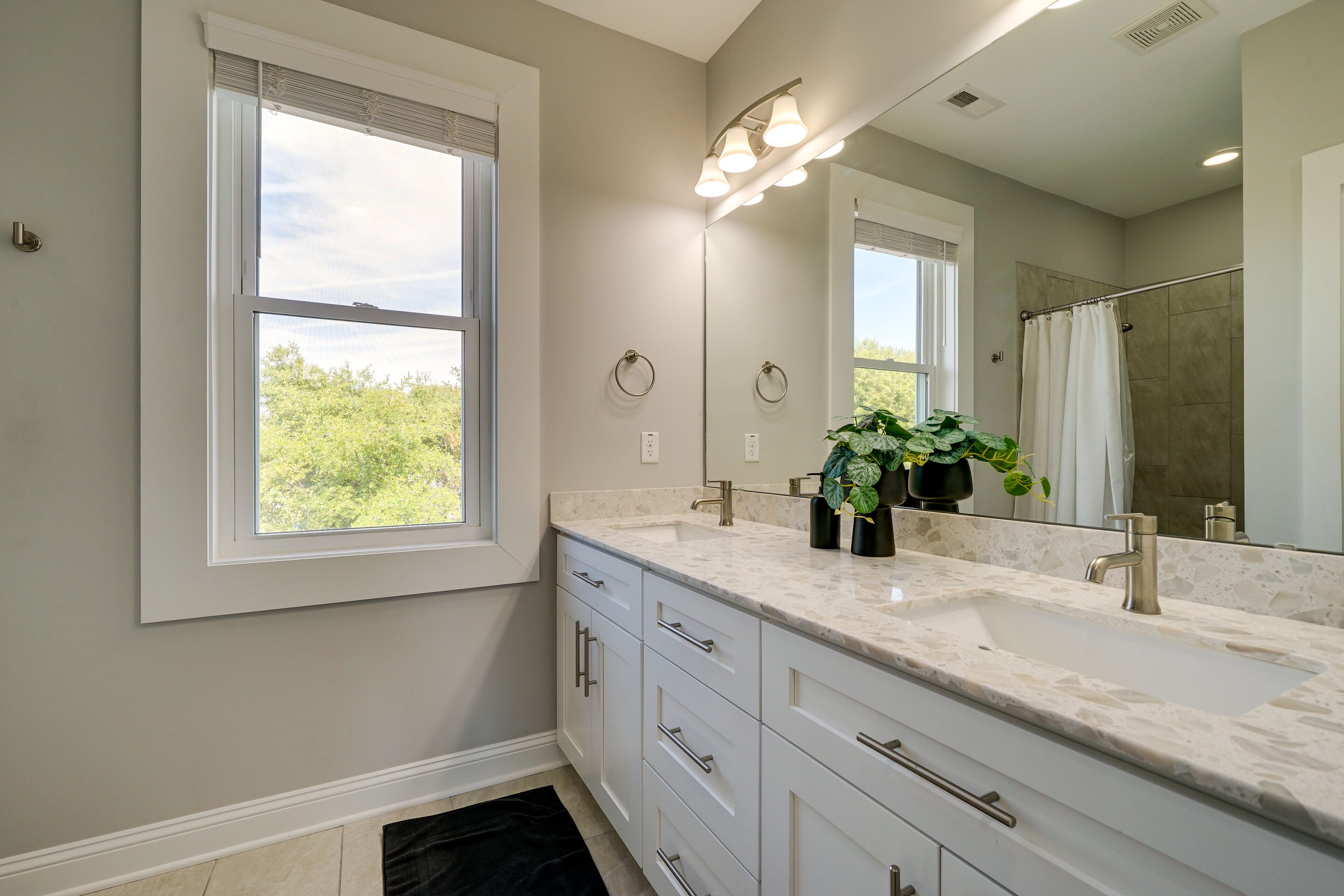 A modern bathroom with a large window, a sink with a marble countertop, and a mirror with lights above it.