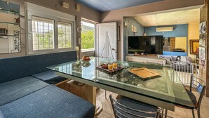 Una cocina moderna con una mesa de comedor de cristal, una zona de estar con un sofá azul y un televisor al fondo.