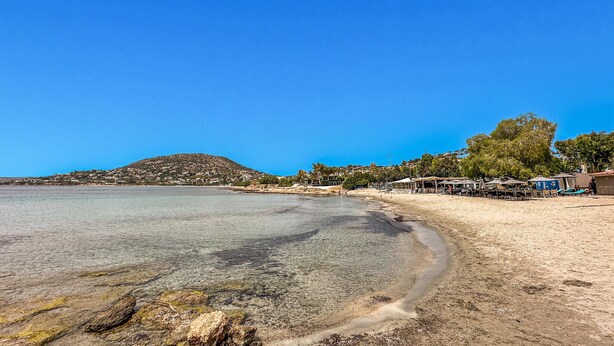 Una playa con agua cristalina, una orilla arenosa y una colina al fondo. Hay edificios y árboles a lo largo de la costa.