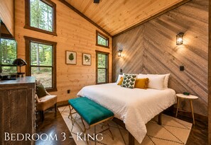A bedroom with a large bed, a bench, a desk, and a chair. There are windows and wall-mounted lights.