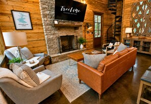 A cozy cabin-style room with a stone fireplace, a mounted television, and a comfortable seating area with a sofa and armchairs.