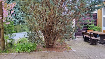 Eine Terrasse mit Backsteinboden, einem Holztisch und Bänken sowie verschiedenen Pflanzen, darunter ein großer Baum und blühende Sträucher.