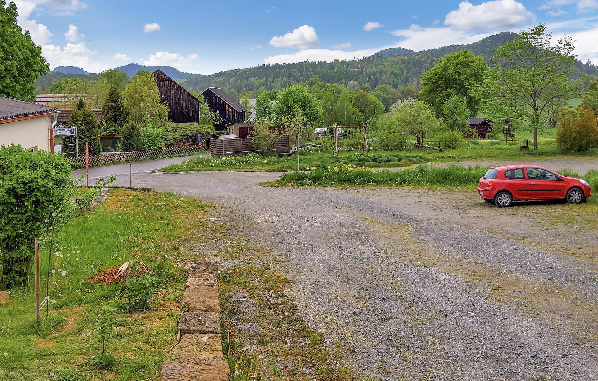 Paisatge rural amb un camí de grava, un cotxe vermell aparcat al costat i un graner de fusta al fons.
