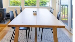 Pitkä, suorakaiteen muotoinen ruokapöytä tuoleineen, suuret ikkunat ja näkymä vehreään maisemaan.