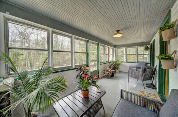A covered porch with large windows, potted plants, and outdoor furniture.
