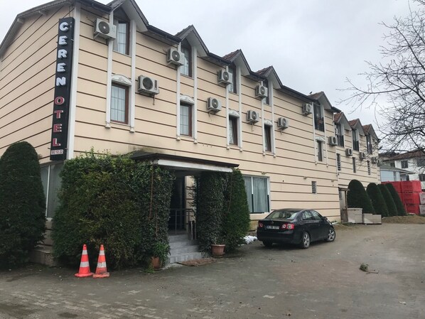 Un hotel con un letrero que dice "CERENOTEL" y un número de teléfono. Se ven unidades de aire acondicionado en el edificio, un coche estacionado adelante y dos conos de tráfico cerca de la entrada.