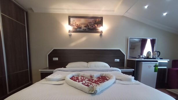 Una cama bien hecha con un arreglo de pétalos de rosa en forma de corazón, un refrigerador pequeño, un escritorio con espejo y un cuadro sobre la cama.