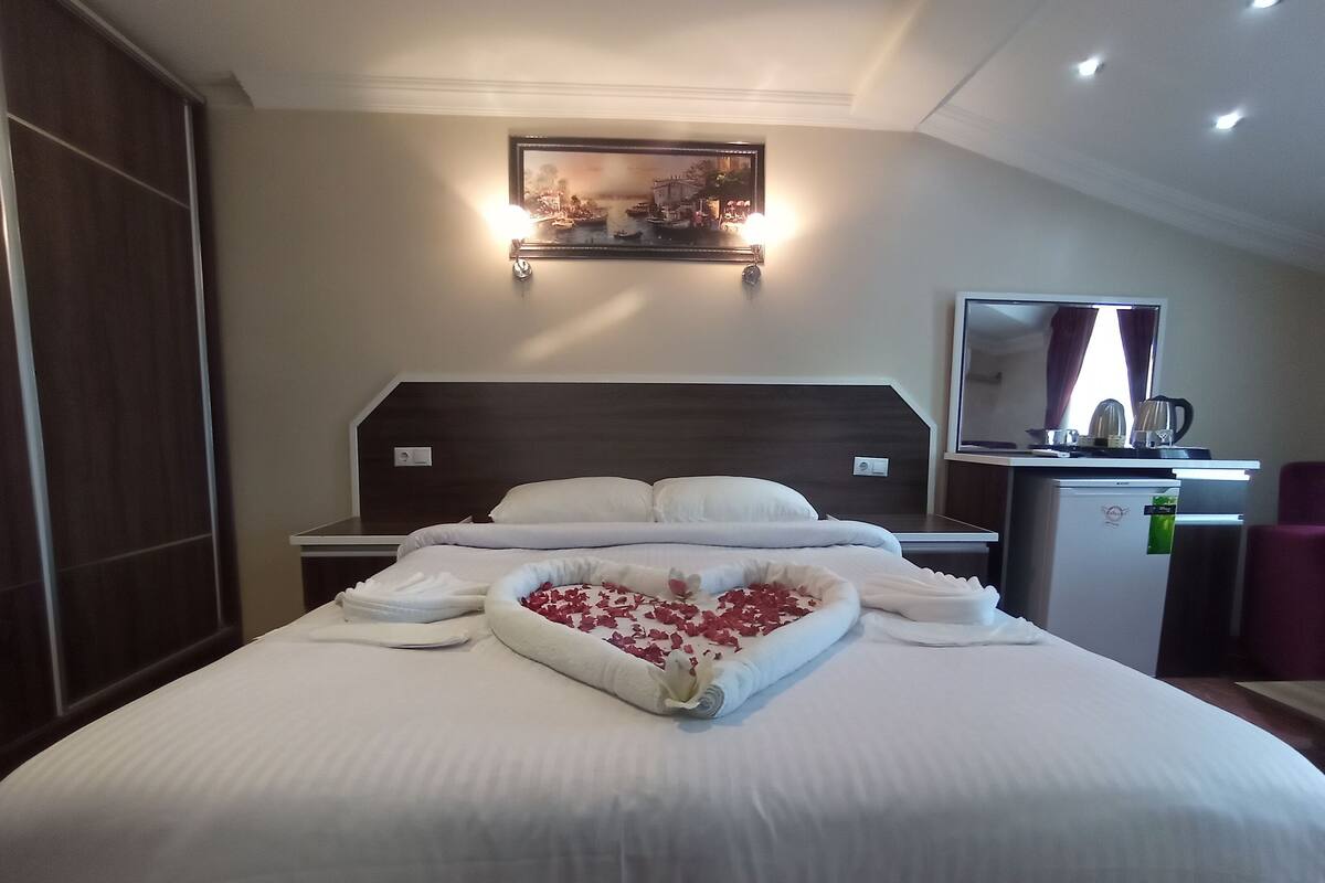A neatly made bed with a heart-shaped arrangement of rose petals in the center, a small refrigerator, a desk with a mirror, and a painting above the bed.