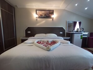 A neatly made bed with a heart-shaped arrangement of rose petals in the center, a small refrigerator, a desk with a mirror, and a painting above the bed. - Ceren Otel (Sapanca)