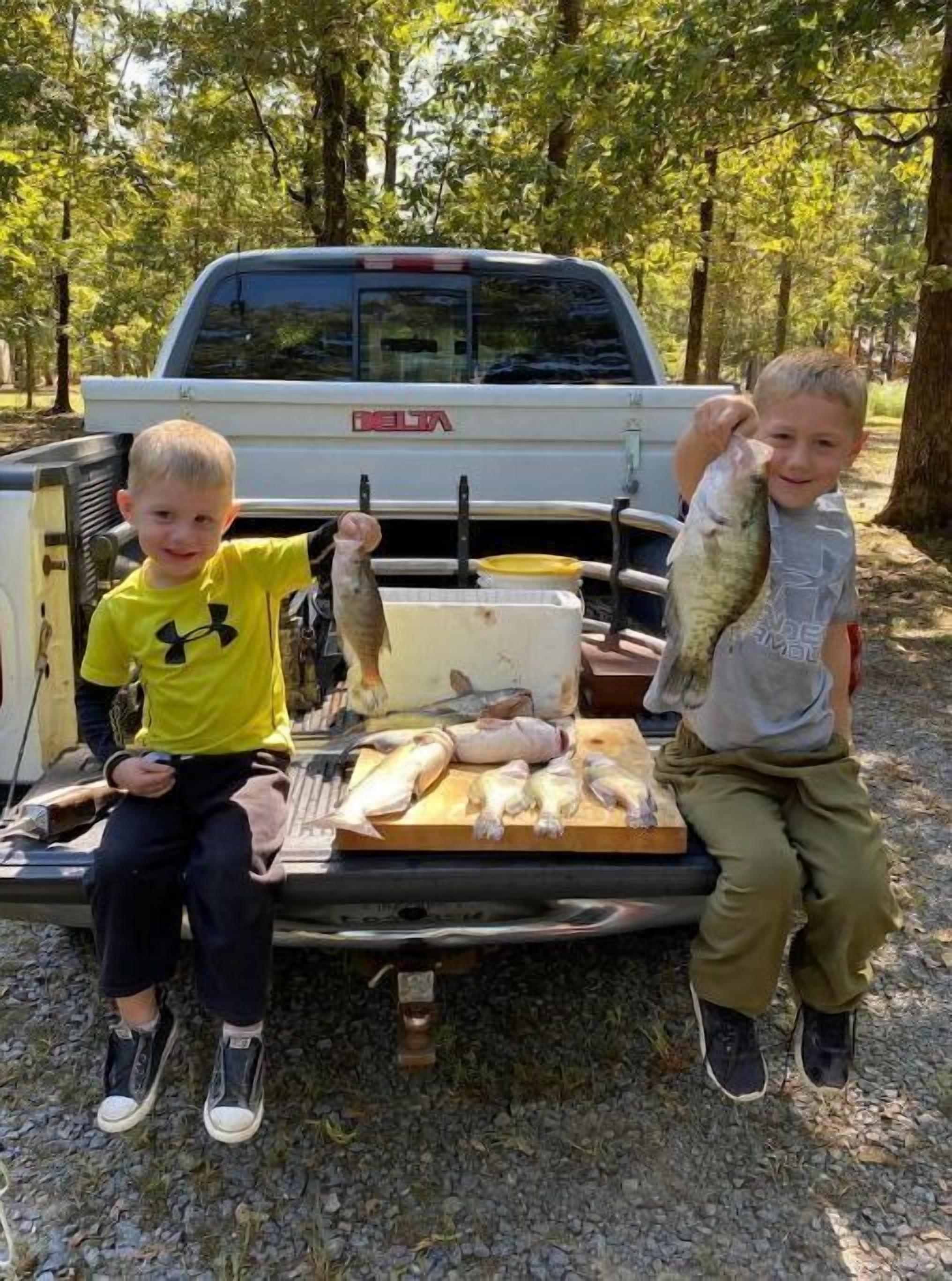 Two children sitting on the back of a truck with a cooler full of fish.