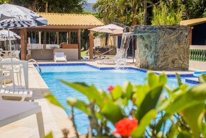 Una piscina con sillas de descanso y sombrillas, una cascada y una jardinera de piedra.