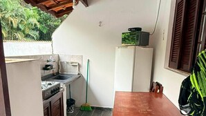 Une petite cuisine équipée d’une cuisinière à gaz, d’un évier et d’un réfrigérateur. On y trouve une table et des chaises en bois, et un balai est appuyé contre le mur.