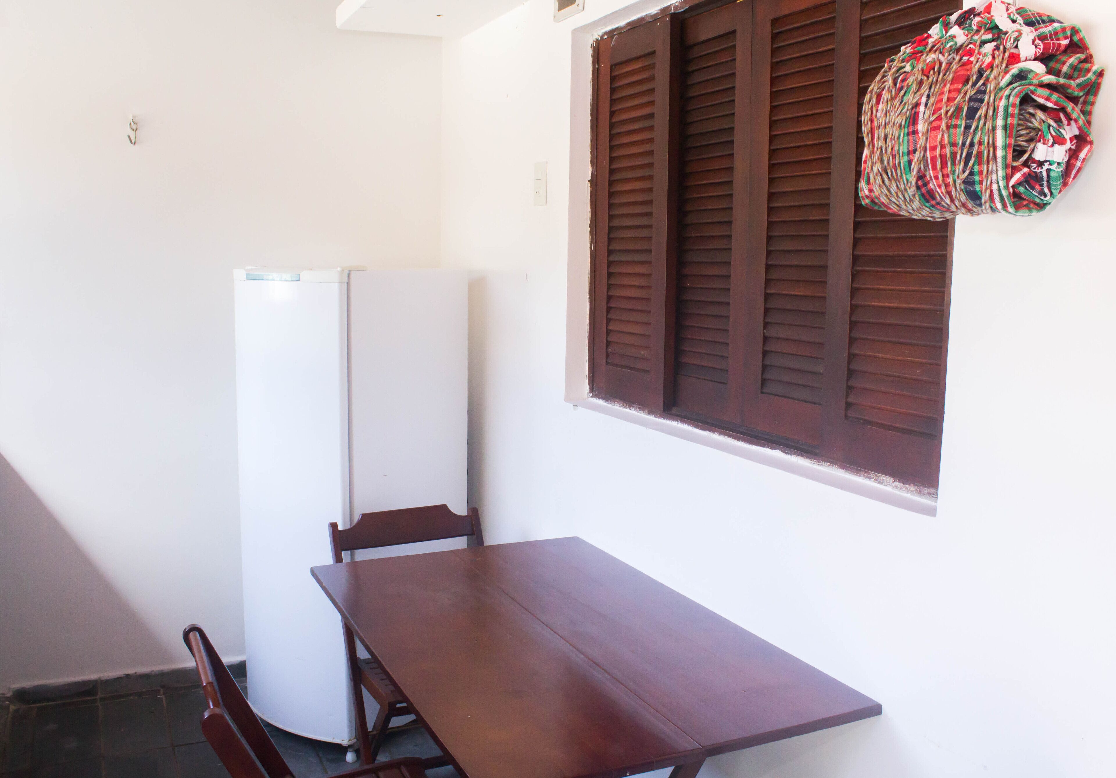 A small room with a wooden table and chairs, a white refrigerator, and a window with brown shutters.