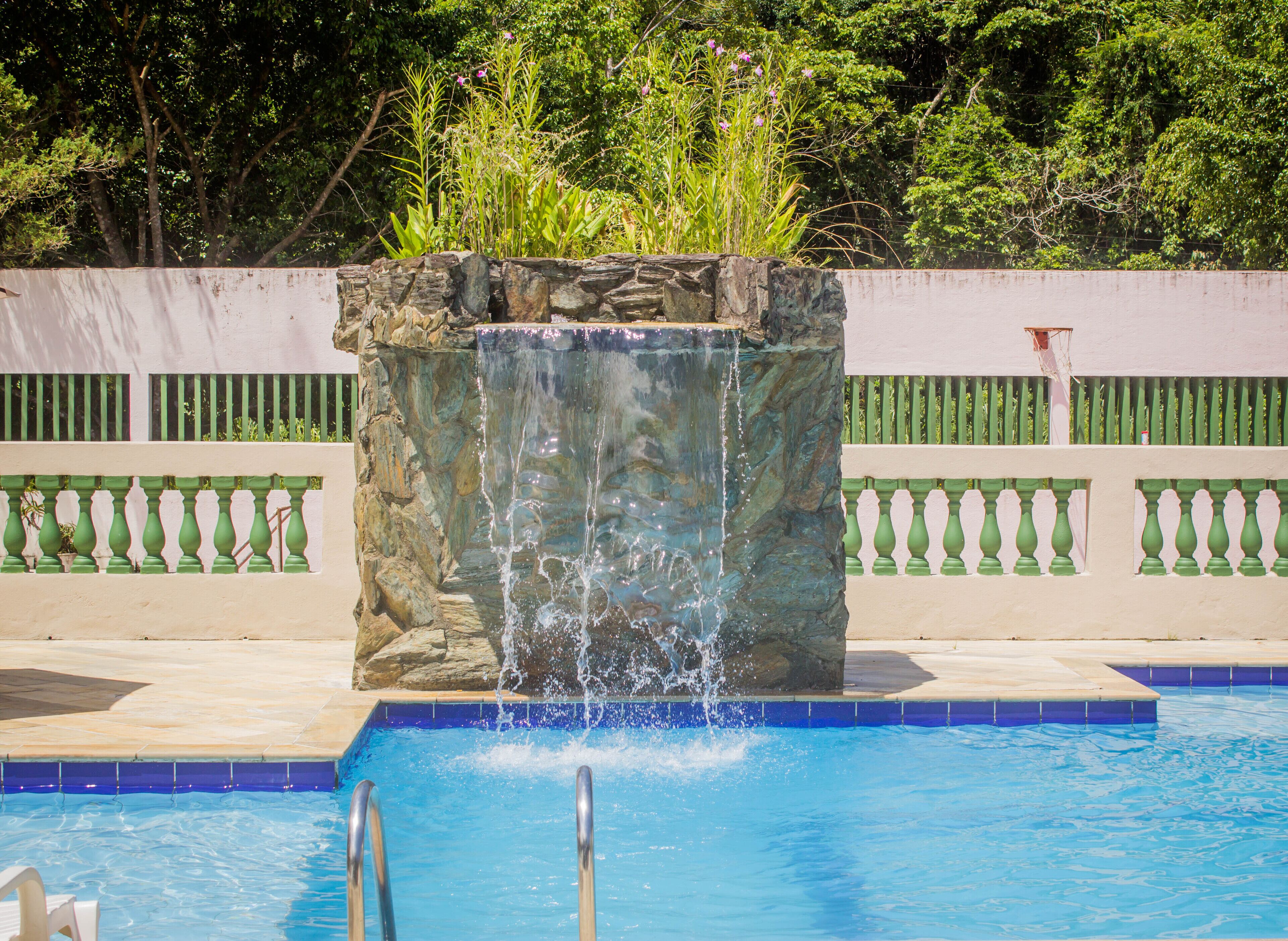 Ein Pool mit einem Wasserfall-Feature aus Stein und einem grünen Zaun im Hintergrund.