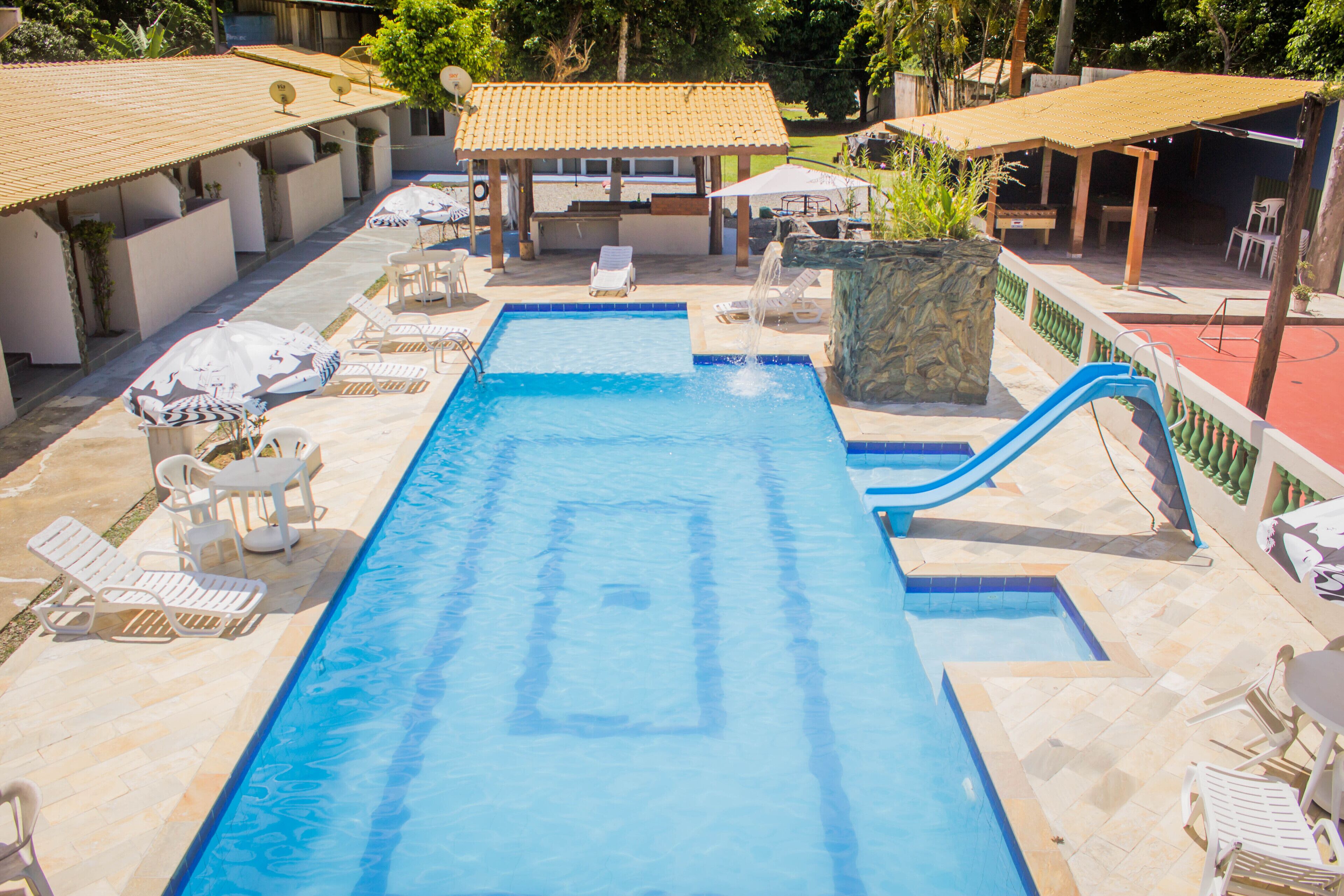 Una piscina con sillas de descanso y sombrillas, rodeada de zonas de estar cubiertas y una jardinera de piedra.