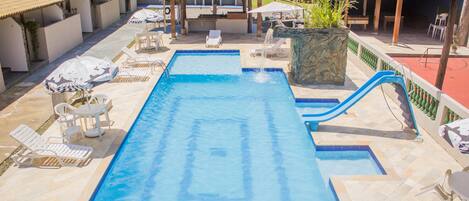 Una piscina con sillas de descanso y sombrillas, rodeada de zonas de estar cubiertas y una jardinera de piedra.