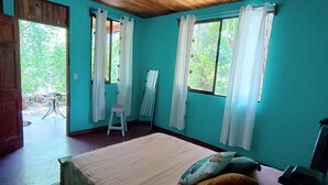 Una habitación con techo de madera, una cama con almohadas, un taburete pequeño y una ventana con cortinas.