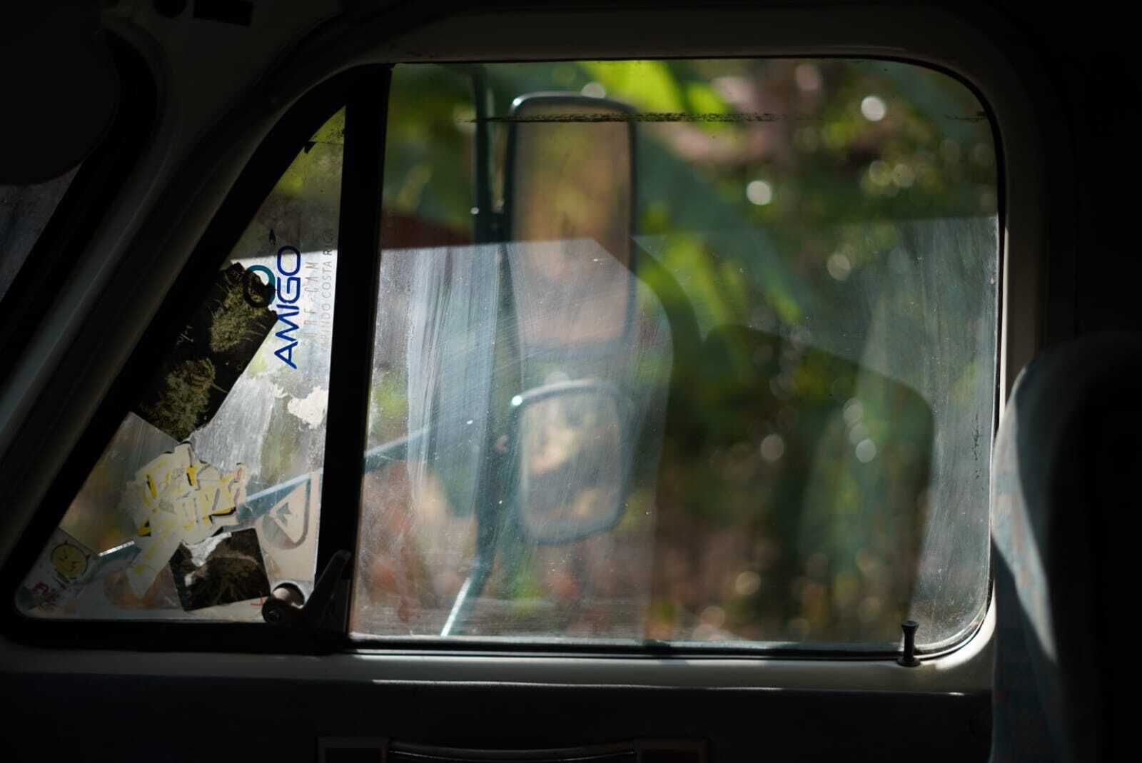 El interior de un coche, con un reflejo en la ventana que muestra a una persona y algo de vegetación al exterior.