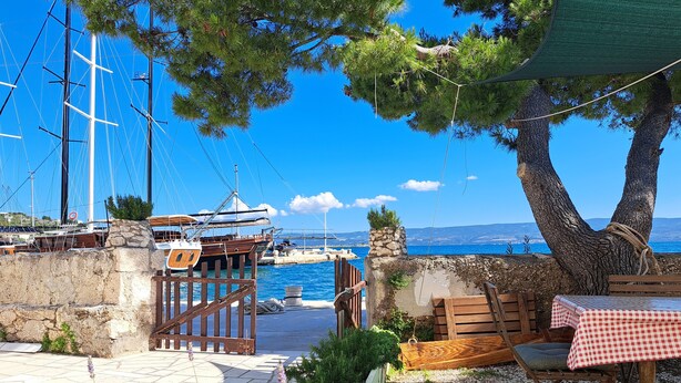 Une terrasse en bord de mer, avec une table et des chaises, un mur en pierre et une vue sur la mer et les bateaux.