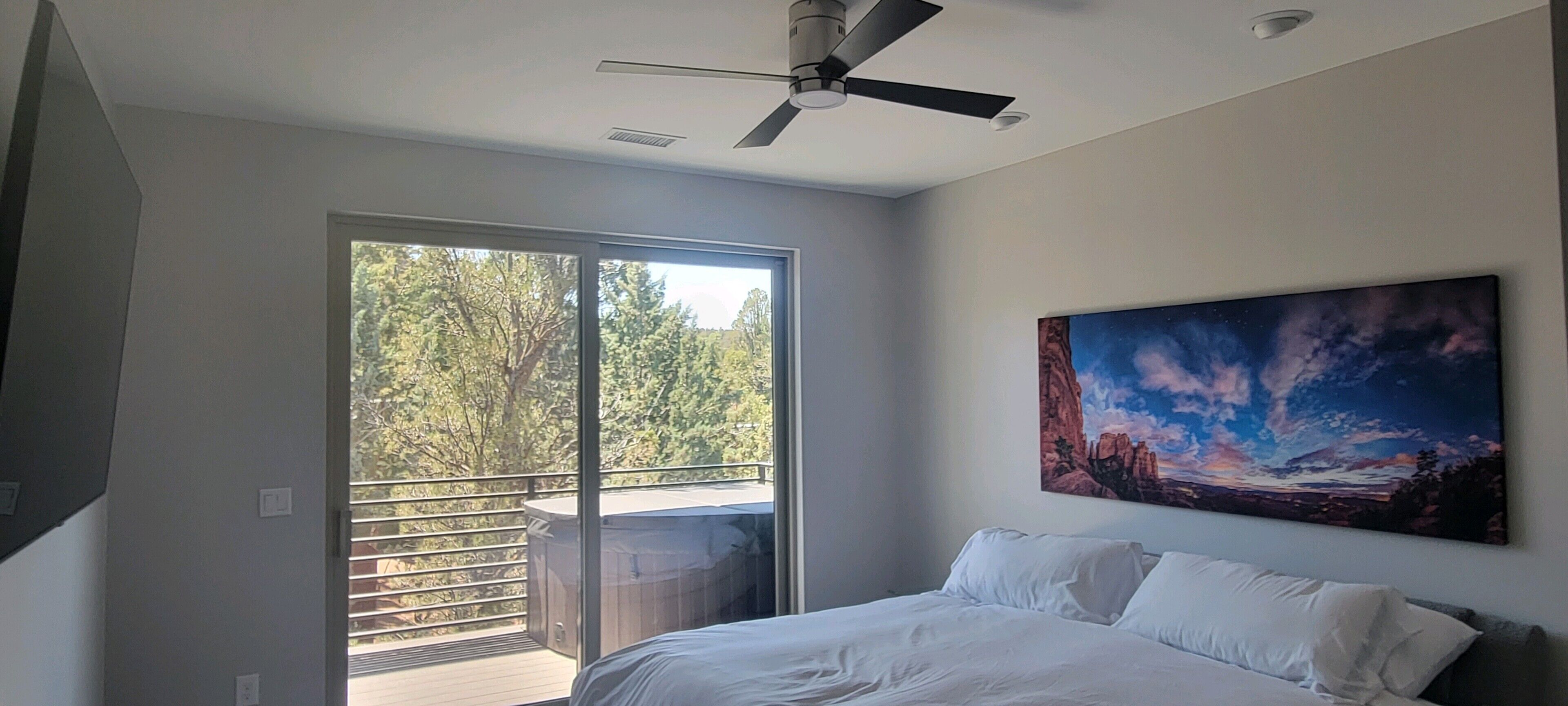 Un dormitorio con cama, ventilador de techo, un cuadro y balcón con vistas a árboles.