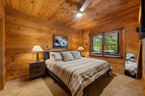 A wooden cabin bedroom with a bed, two bedside tables, a ceiling fan, a window with blinds, and a framed picture on the wall.
