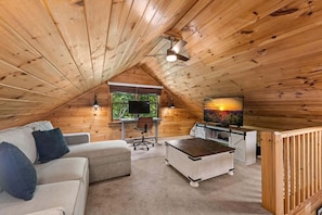 A cozy attic room with a wooden ceiling, a white desk with a computer, a television, a cushioned sofa, and a small wooden table. - Copper Ridge Hideaway - Dog Friendly | Pac Man Arcade Game | Hot Tub (Mineral Bluff)