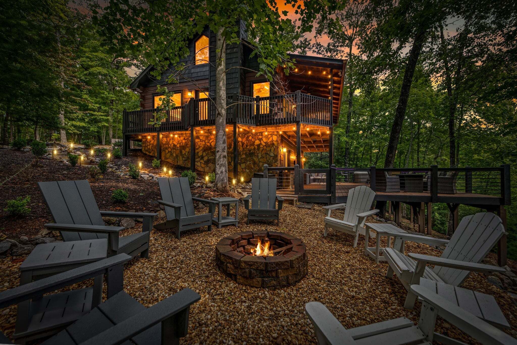 A cabin with a stone exterior, multiple decks, and outdoor seating with Adirondack chairs and a fire pit.