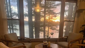 A cozy room with a large window overlooking a forest and a lake, featuring wooden furniture and a small table with a game board. - Waterfront Paradise! Boat available for rent. (Waupaca)