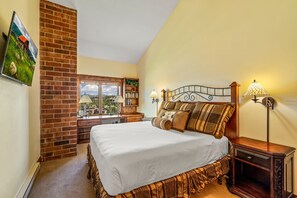 A bedroom with a brick wall, a large bed, a nightstand, a lamp, and a window with a view. - Bear Claw 614 Premium On Mountain Condo with Ski-In/Ski-Out Access! (Steamboat Springs)
