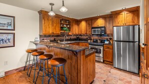 A kitchen with wooden cabinets, a stainless steel refrigerator, a microwave, a sink, and a bar stool area. - Bear Claw 614 Premium On Mountain Condo with Ski-In/Ski-Out Access! (Steamboat Springs)