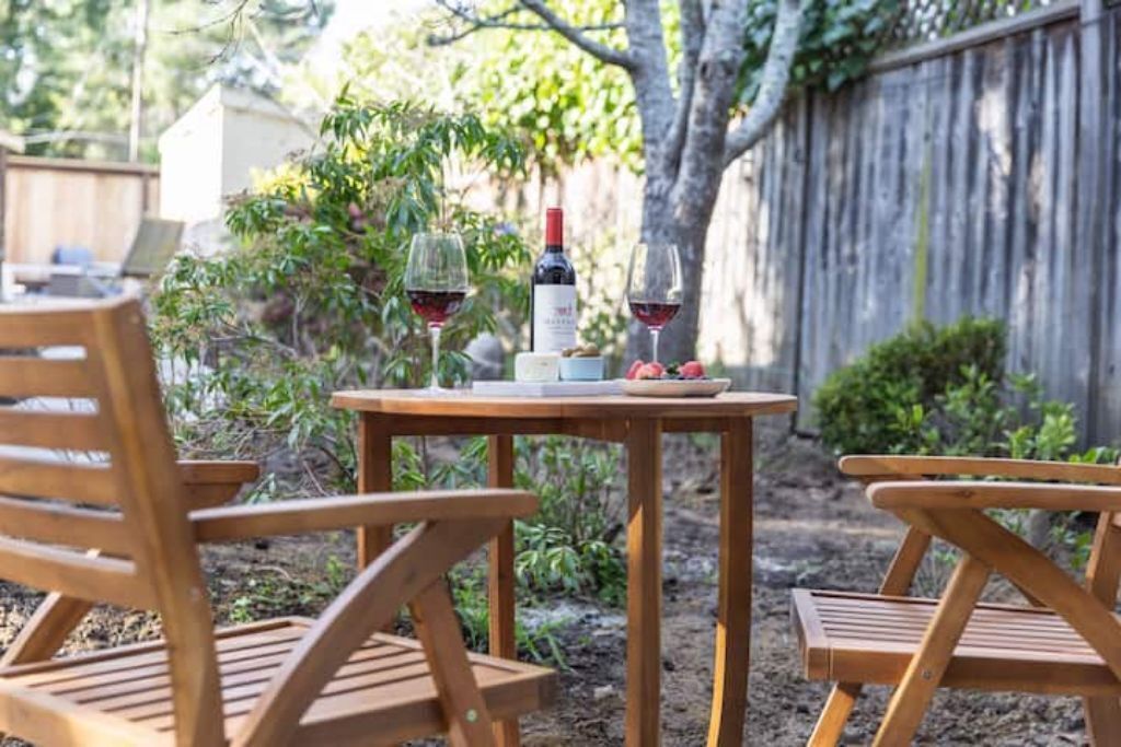 Une table en bois placée à l’extérieur, avec deux chaises, une bouteille de vin et un verre de vin rouge dessus.