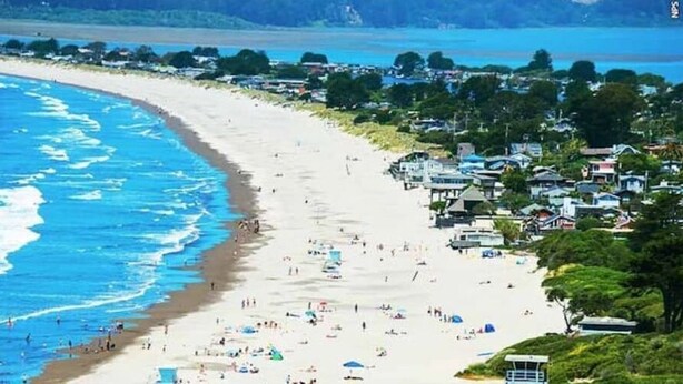 Una playa con una larga extensión de arena, algunos sombrillas y unas pocas personas. Se ven edificios a lo largo de la playa y una torre de salvavidas.