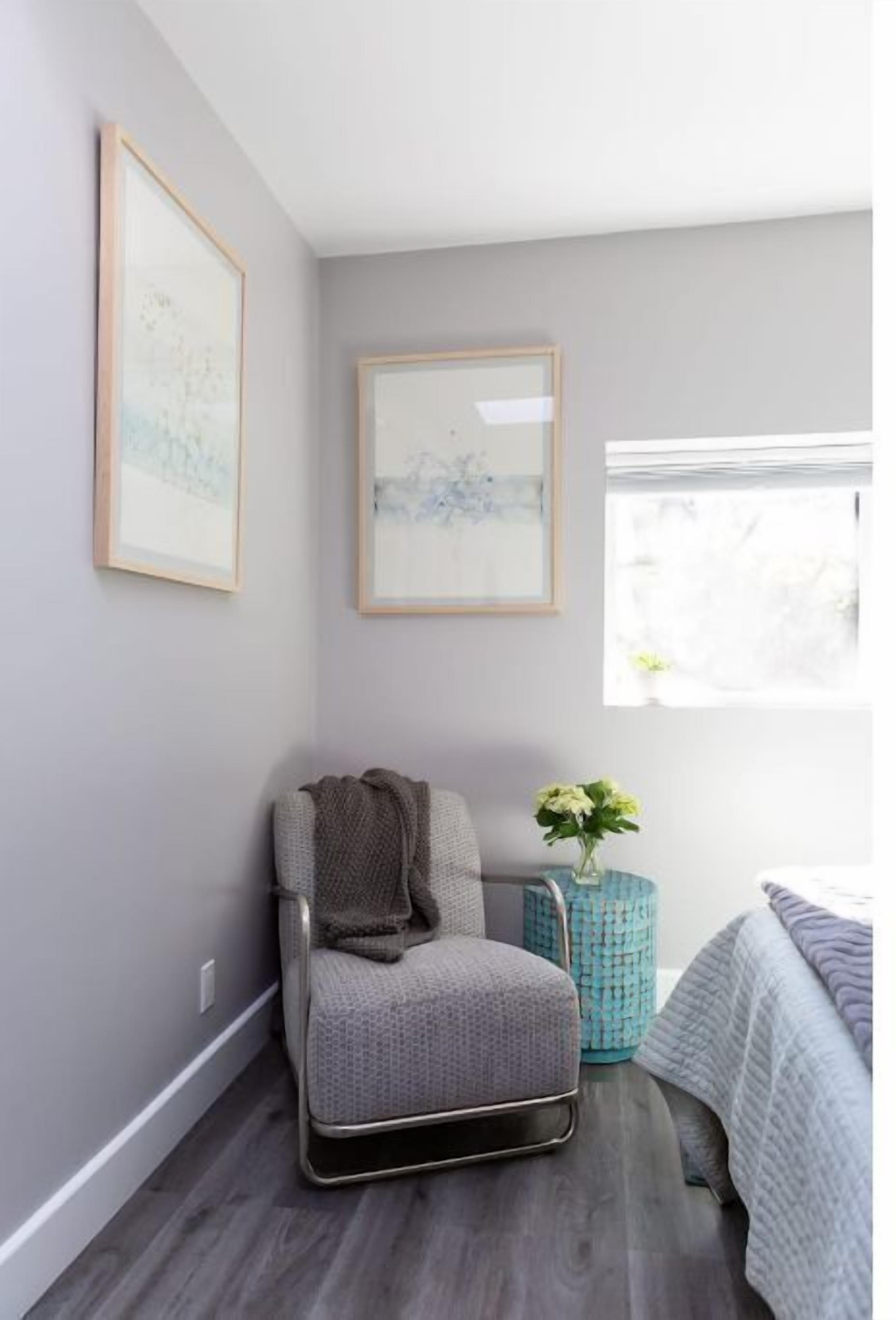 Een kamer met een grijze stoel, een klein tafeltje met een vaas en twee ingelijste foto's aan de muur.