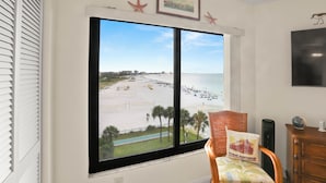 A room with a large window overlooking a beach, a wicker chair, a wooden cabinet, and a framed picture on the wall. - 🌴 Caprice Resort Unit #507 – Gulf Front Stunner with Full Kitchen & Balcony Views (Saint Pete Beach)