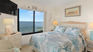 A bedroom with a large window, a bed with a starfish and coral pattern, a television, and a painting on the wall. - 🌴 Caprice Resort Unit #507 – Gulf Front Stunner with Full Kitchen & Balcony Views (Saint Pete Beach)