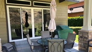 Um pátio externo com mesa de vidro rodeada por cadeiras, churrasqueira coberta e porta de correr que dá para um jardim.
