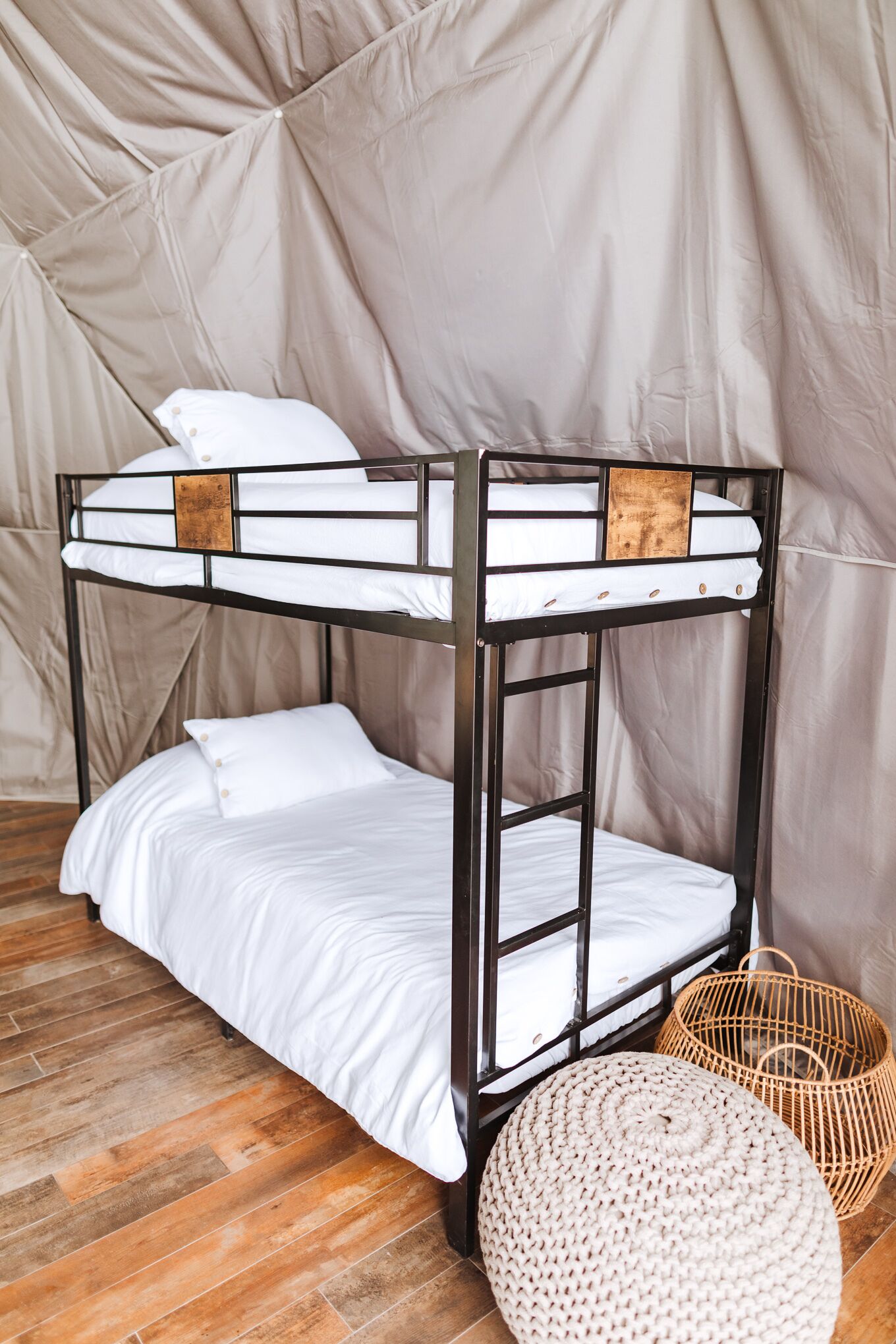 A bunk bed with white bedding and a wooden headboard, a woven basket, and a round ottoman on a wooden floor.