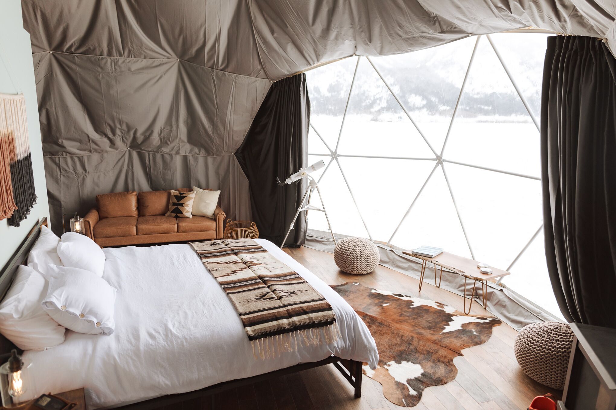 A cozy bedroom with a bed, a sofa, a cowhide rug, and a large window offering a view of a snowy landscape.