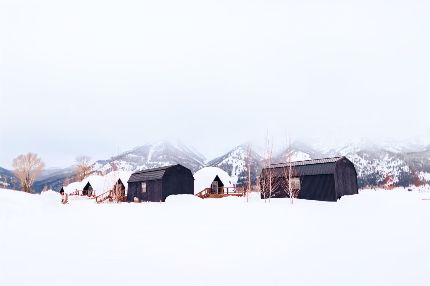 A snow-covered landscape with three barns and a mountain in the background.