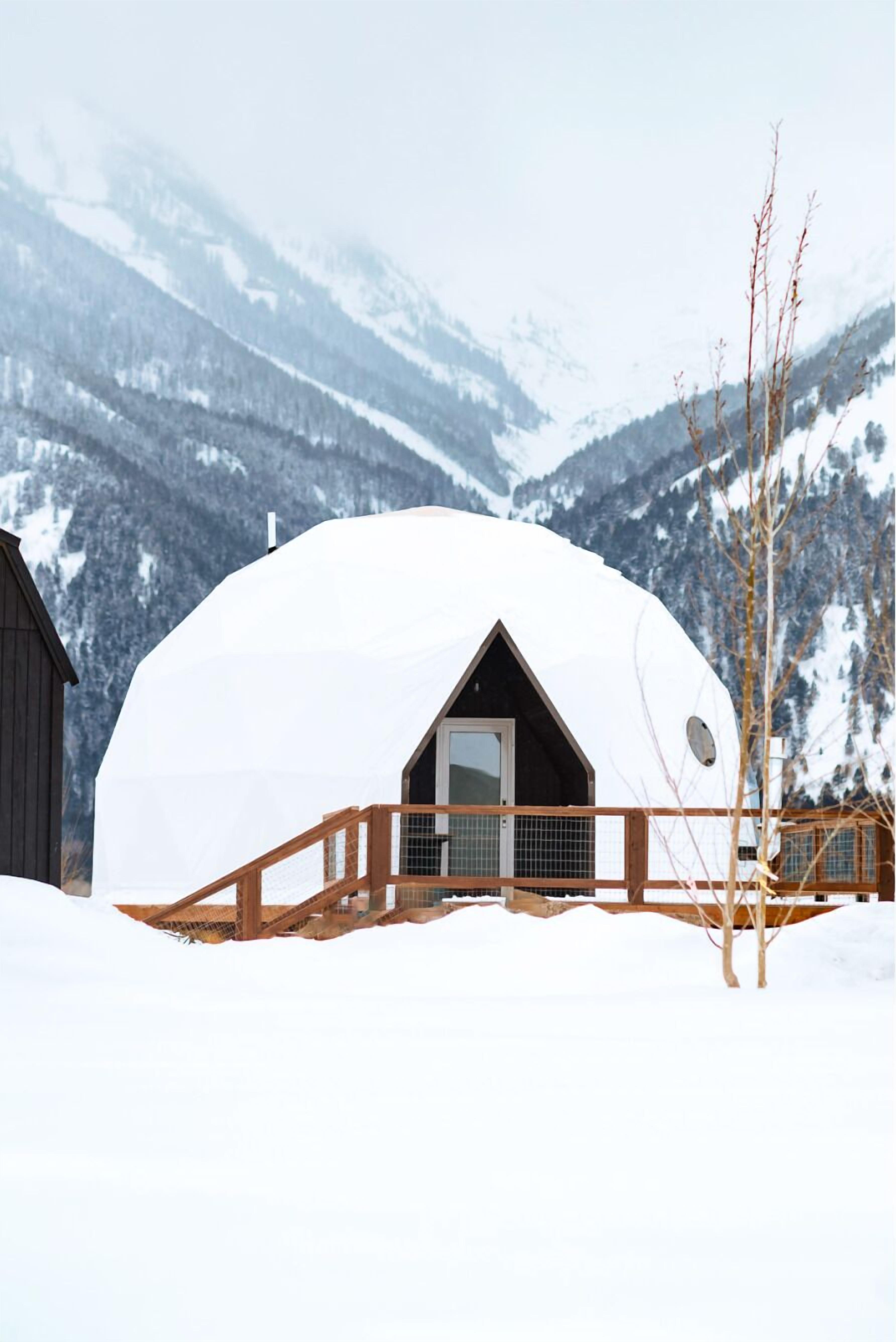 A snow-covered dome structure with a wooden deck in front, set against a mountainous backdrop.