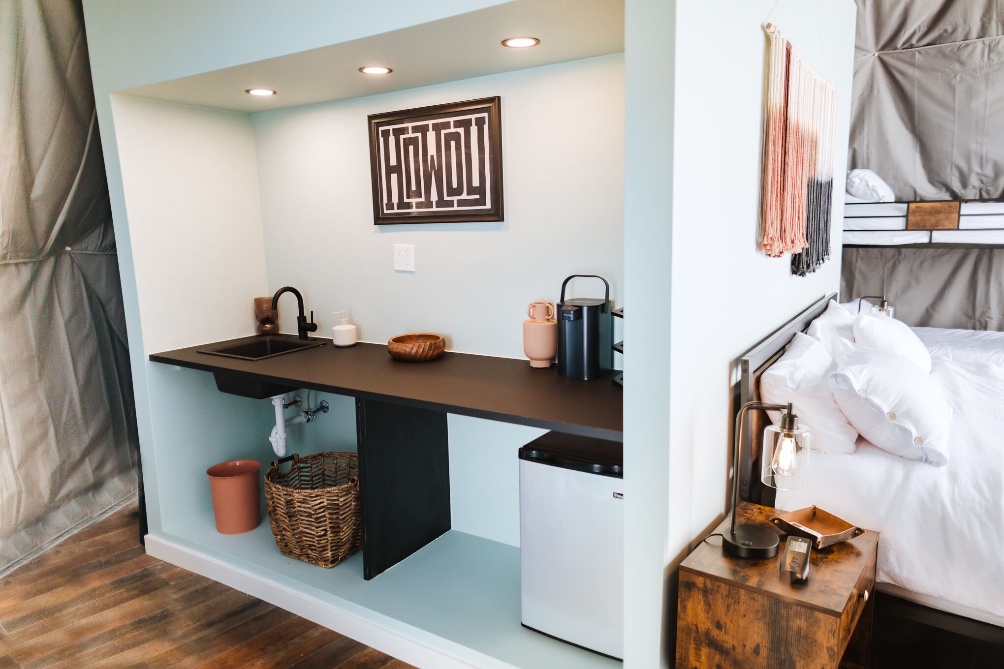 A compact kitchenette with a sink, a small refrigerator, and a coffee maker. There is a bed with a white comforter and a bedside table with a lamp.